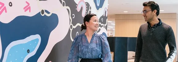 A man and women walking and talking in the workplace, in front of a colourful Aboriginal painting.