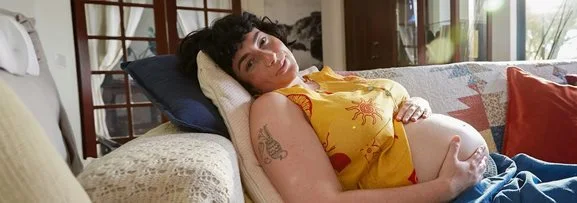 A pregnant women laying on a couch holding her belly and smiling at the camera.