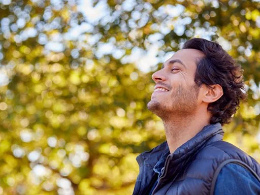 A man standing outside in nature feeling content and happy, smiling and looking to the sky.