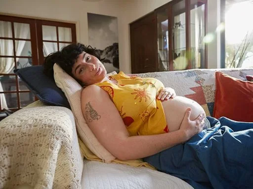 A pregnant women laying on a couch holding her belly and smiling at the camera.