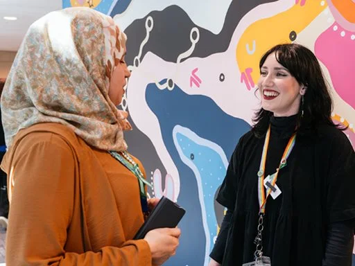 Two women smiling and talking in the workplace, in front of an Aboriginal painting.