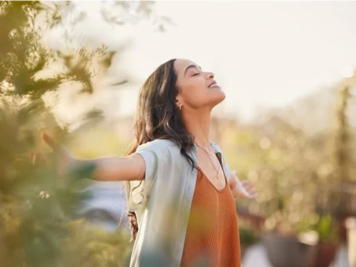 A women standing outside in nature feeling content and happy hold her hands out, smiling and looking to the sky.