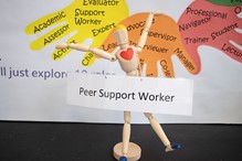 Image of a wooden doll holding a sign saying Peer Support Worker