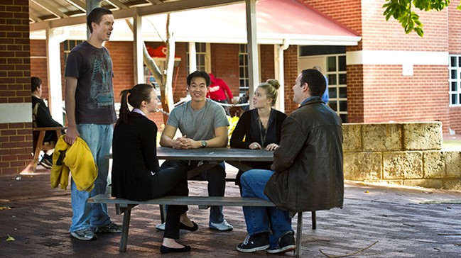Five young people are gathered around a picnic table in the courtyard of a building.